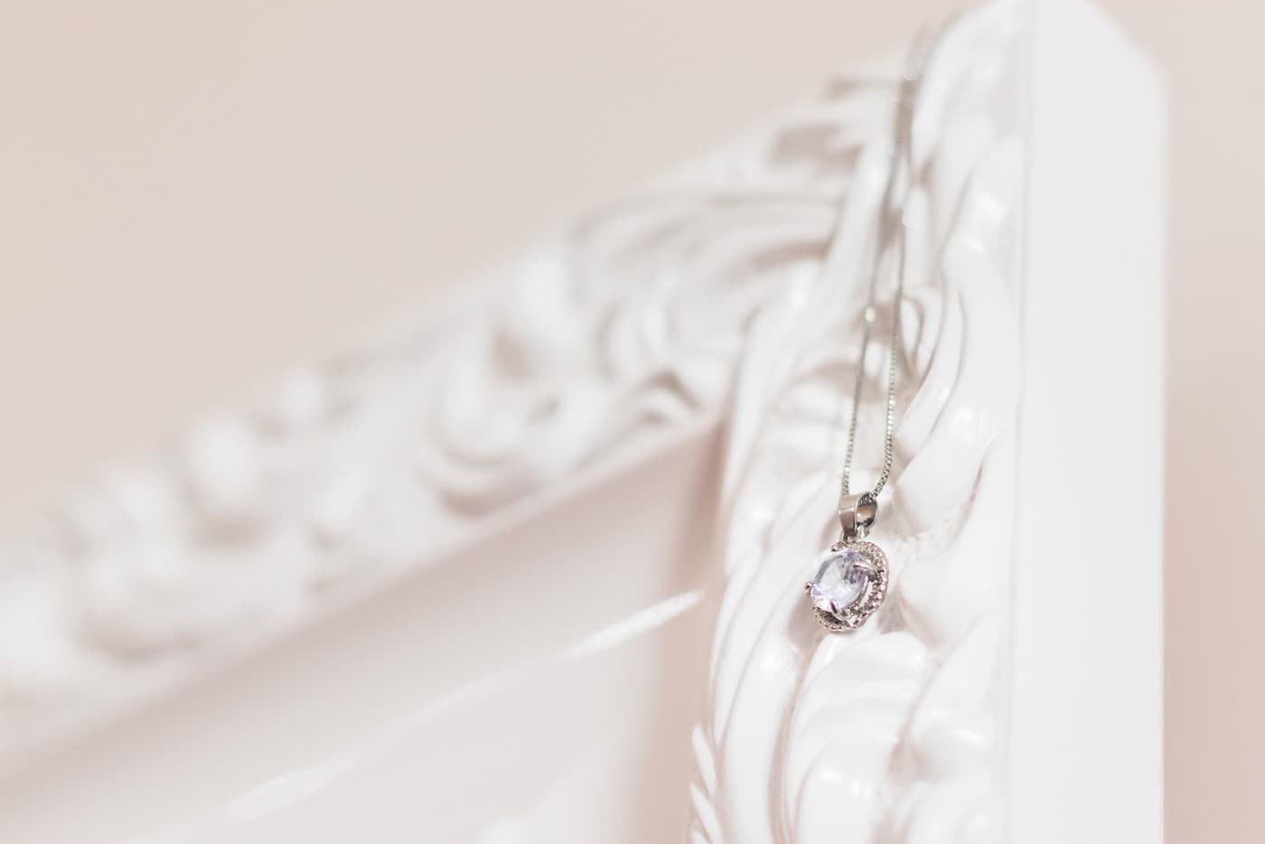 A silver pendant necklace with a small clear gemstone, hanging against a carved white frame