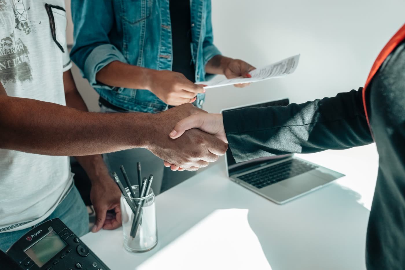 Two people shaking hands over a desk with documents and a laptop visible